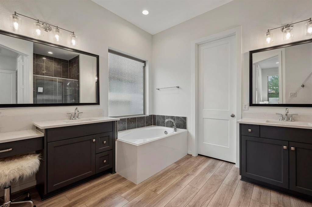 This bathroom features a modern design with dual vanities, sleek black cabinetry, and chic lighting. It includes a spacious bathtub, a large window for natural light, and a separate shower with dark tile accents. The wooden floor adds warmth to the contemporary style.