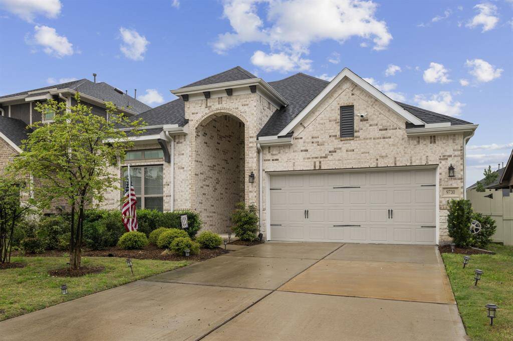 This photo features a modern, single-story brick home with an arched entryway and a two-car garage. The well-maintained front yard includes lush landscaping and a pathway leading to the entrance. The house has a neat, inviting curb appeal.