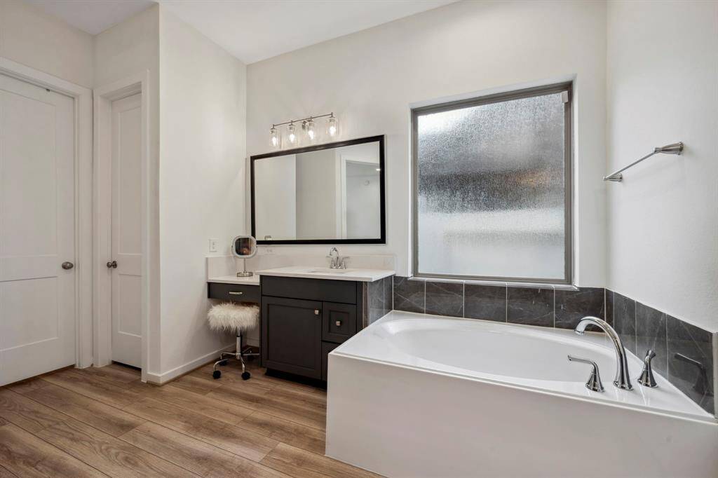 This modern bathroom features a sleek design with a large soaking tub, a stylish vanity with a mirror and lighting, and wood-look flooring. It offers a bright and inviting space with a frosted window for privacy.