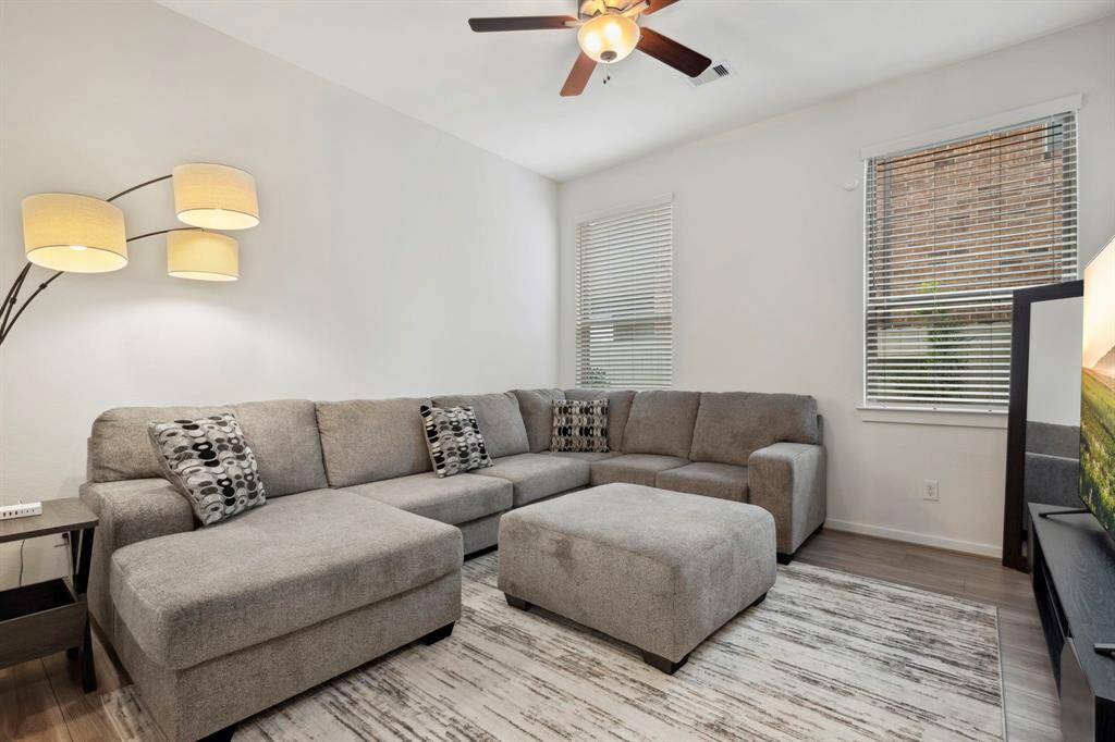 This cozy living room features a modern gray sectional sofa with patterned cushions, a stylish floor lamp, and a ceiling fan. Natural light filters in through the windows, highlighting the sleek, light-colored flooring. Perfect for relaxation and entertainment.