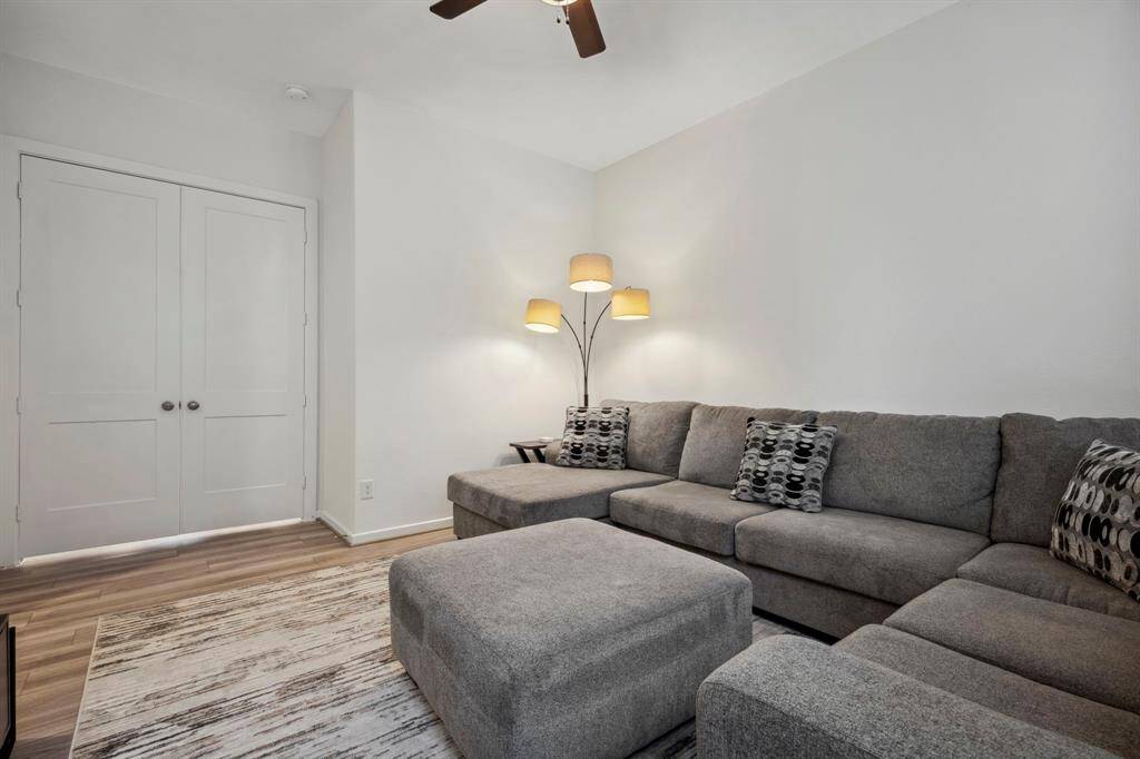 Cozy living room with a large gray sectional sofa, stylish pillows, and a modern floor lamp. The space features light wood flooring, a neutral area rug, and a ceiling fan. Double doors add charm and functionality.