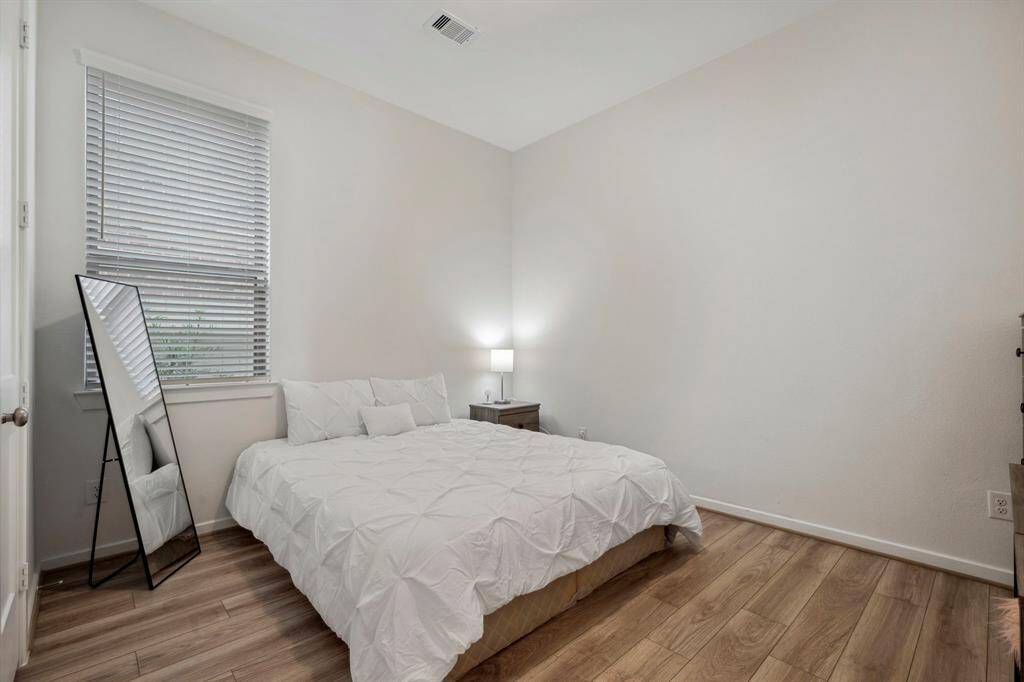 This is a cozy, minimalist bedroom with wood flooring, featuring a comfortable bed, a bedside table with a lamp, a large window with blinds, and a full-length mirror.