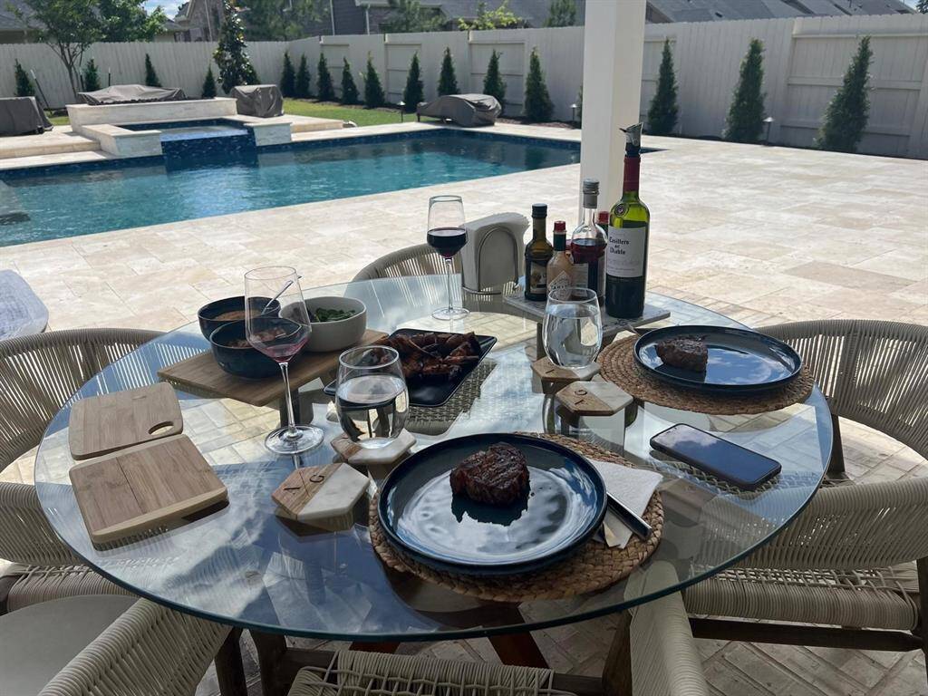 This photo showcases a stylish outdoor dining area with a glass table set for a meal, including wine and steak. The space overlooks a luxurious pool and spa, surrounded by a well-maintained patio and privacy fence, creating an inviting atmosphere for relaxation and entertainment.