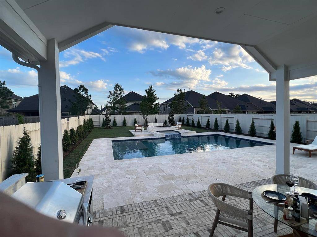 This photo showcases a modern backyard with a spacious pool, bordered by a well-maintained lawn and privacy fence. The covered patio features outdoor dining furniture and a built-in grill, perfect for entertaining.