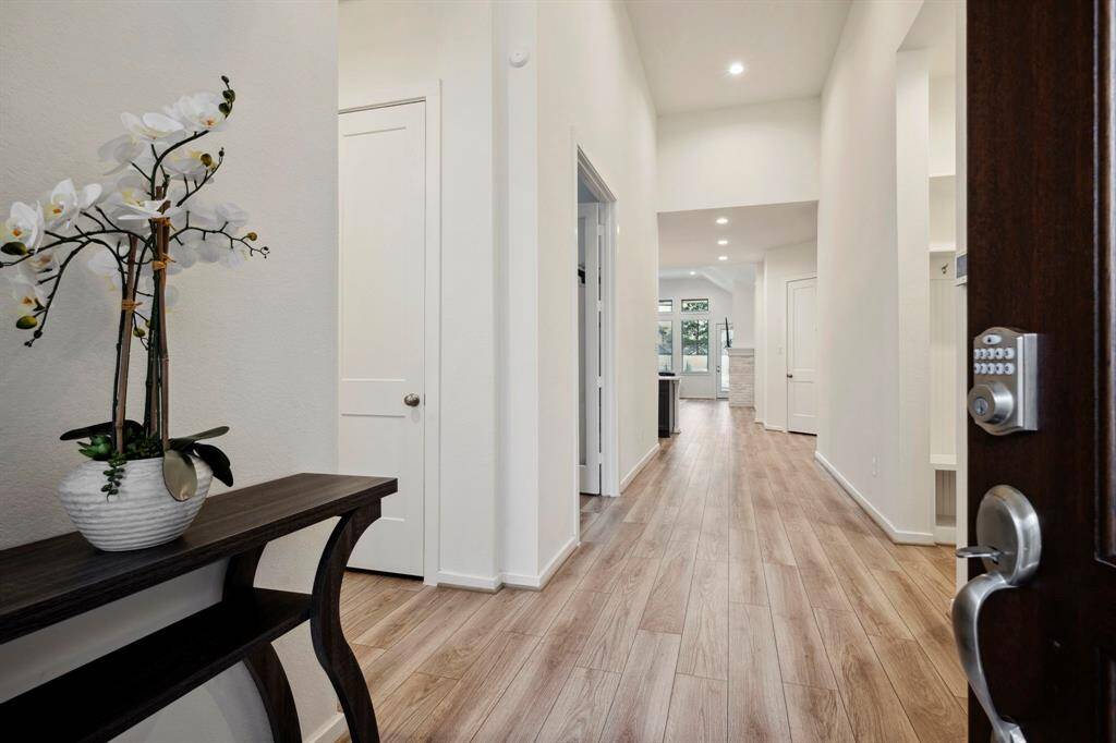 Bright and welcoming entryway with wood floors, white walls, and recessed lighting. A decorative table with an orchid adds a touch of elegance. The hallway leads to an open living space with large windows.