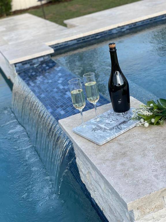 This photo showcases a luxurious pool area with a waterfall feature, elegant stonework, and a relaxing ambiance enhanced by a bottle of champagne and glasses, perfect for outdoor entertaining or unwinding.