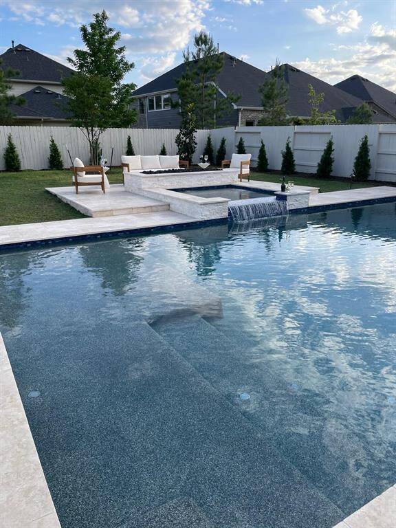 This backyard features a luxurious pool with a modern waterfall feature, surrounded by a spacious patio area with seating. The fenced yard offers privacy, complemented by well-maintained landscaping and trees, creating an inviting outdoor oasis.