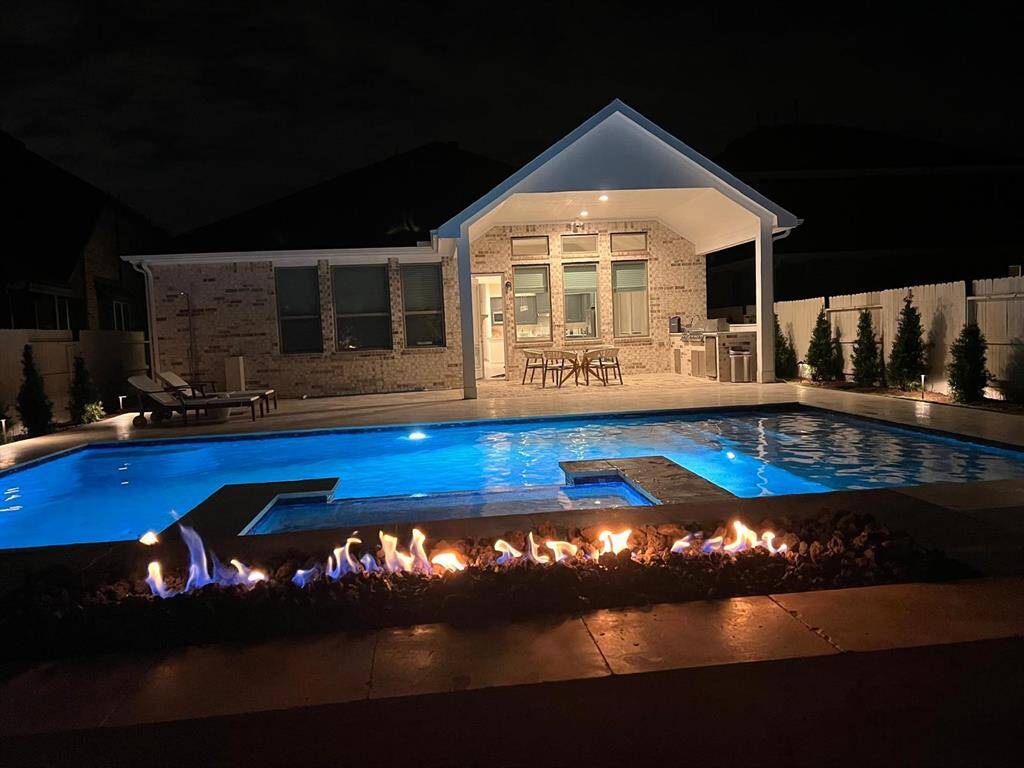 This photo showcases a beautifully lit backyard at night, featuring a luxurious pool with a hot tub and an inviting fire pit in the foreground. The pool area is complemented by a covered patio with outdoor seating and a kitchen, perfect for entertaining.