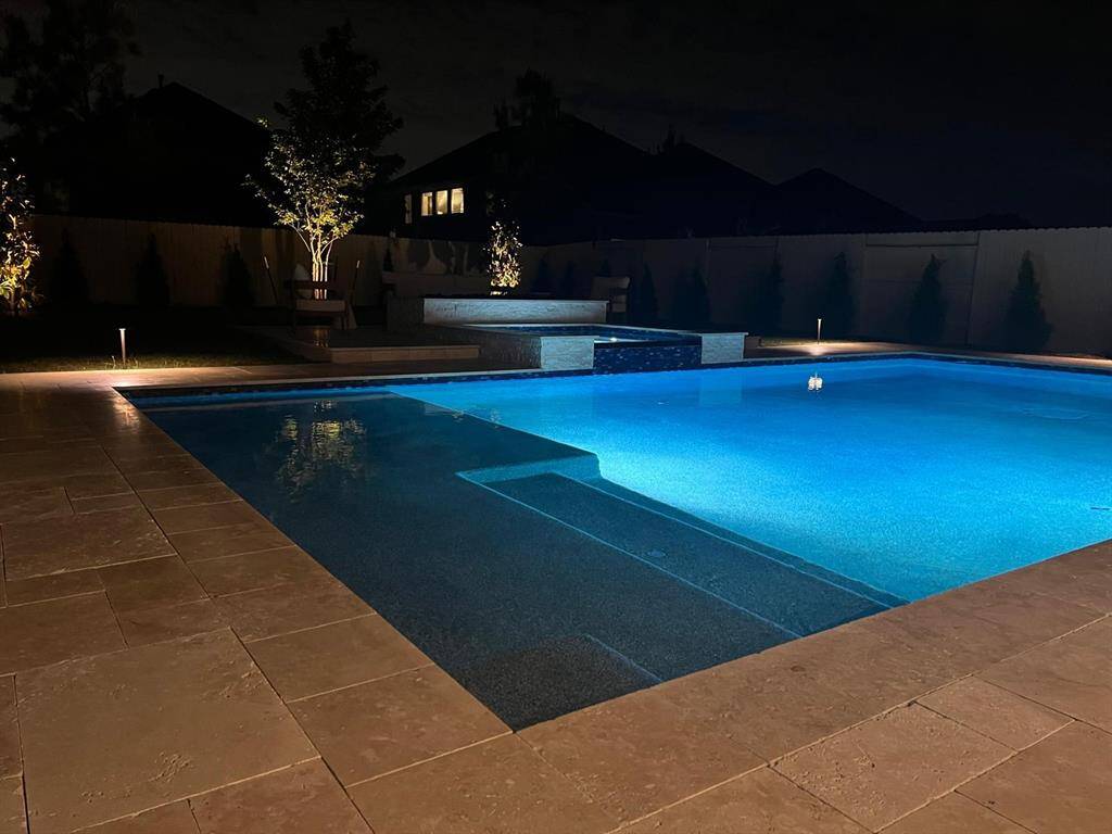 This photo showcases a beautifully lit backyard at night, featuring a spacious swimming pool with a modern design. The surrounding area has tasteful landscaping and ambient lighting, creating an inviting and serene outdoor space.