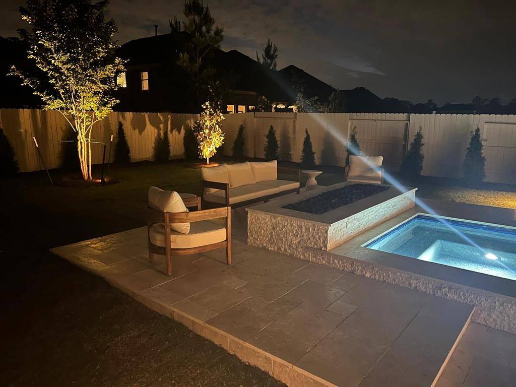 This photo showcases a cozy, well-lit backyard during nighttime, featuring a small pool with surrounding patio seating. The area is enclosed by a wooden fence and enhanced with soft landscape lighting, creating a welcoming outdoor space.