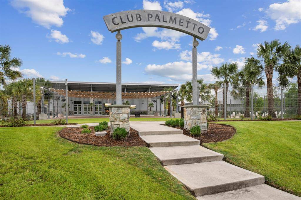 This photo showcases "Club Palmetto," featuring a welcoming entrance with a stone pathway, lush green landscaping, and palm trees. The club building in the background suggests amenities and a community-centered environment, ideal for relaxation and social activities.