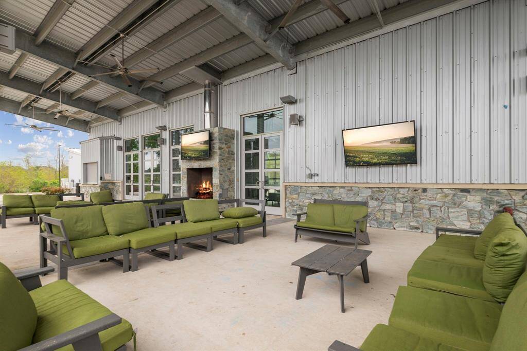 This outdoor living space features a covered patio with a metal roof, green cushioned seating, a stone fireplace, and two mounted TVs, offering a comfortable and stylish area for relaxation and entertainment.
