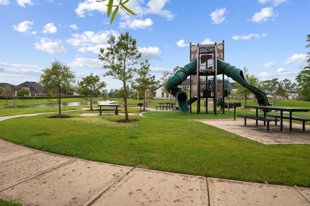 This photo showcases a well-maintained community park featuring a modern playground with slides, picnic tables, and open green spaces. Ideal for families, the area is surrounded by trees and has a picturesque pond view, offering a serene and inviting environment.