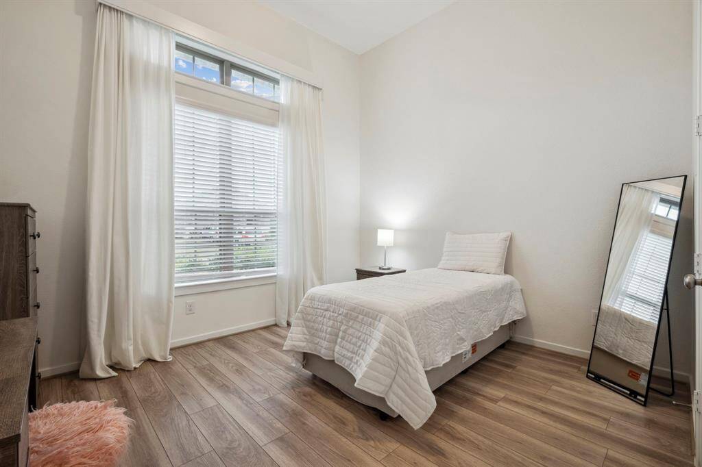 This cozy bedroom features a single bed with a white quilt, a large window with blinds and curtains for ample natural light, a floor mirror, and wood-style flooring, creating a bright and inviting atmosphere.