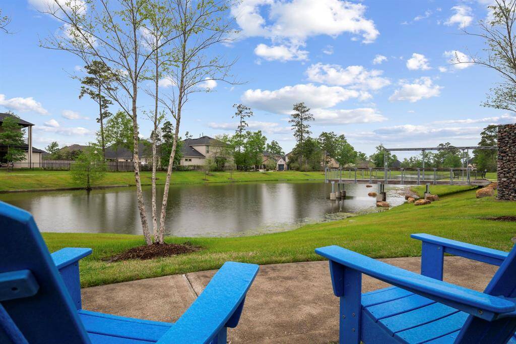 This photo showcases a serene backyard view featuring a tranquil pond and lush greenery, with a charming bridge in the distance. Two blue Adirondack chairs on a patio offer a perfect spot for relaxation.