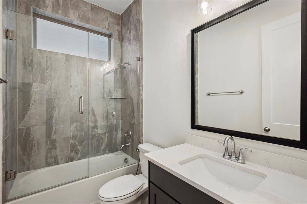 This modern bathroom features a sleek design with a glass-enclosed shower/tub combo, large wall tiles, a white vanity with a black-framed mirror, and contemporary fixtures.