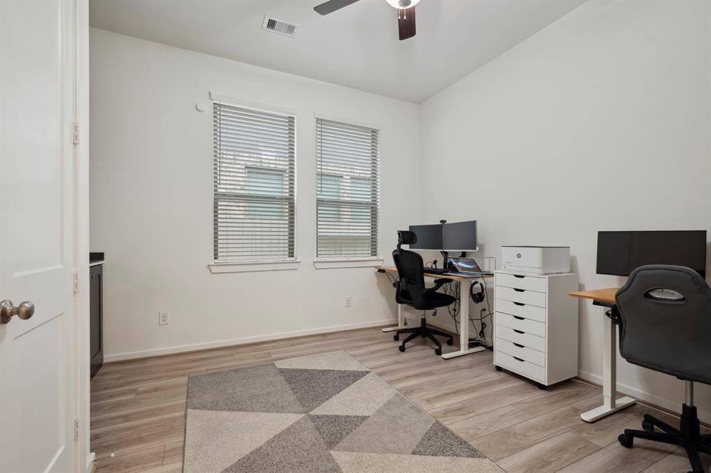 This is a bright, well-organized home office with two desks, modern fixtures, and wood-look flooring. The neutral color palette and ample natural light from the window create a functional and inviting workspace.