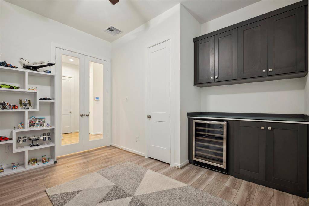 Bright, modern room with sleek dark cabinetry and a wine fridge. Features light wood flooring, built-in shelving, and glass-paneled double doors, offering a stylish and functional space.
