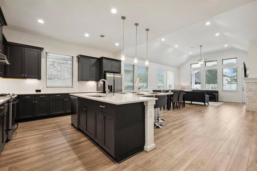 This open-concept living space features a modern kitchen with dark cabinetry, a large island, and pendant lighting. It seamlessly flows into a dining and living area with high ceilings, ample natural light, and a cozy fireplace, all set on elegant wood flooring.
