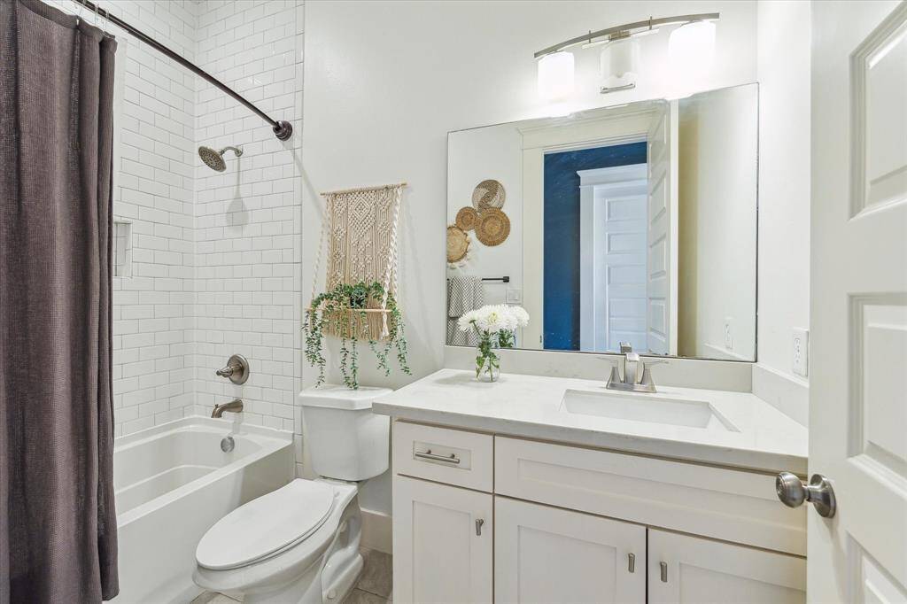 This secondary bathroom features a clean, modern design with white subway tiles, a shower-tub combo, and a sleek vanity with ample counter space. It includes a large mirror, contemporary lighting, and decorative accents for a cozy touch.