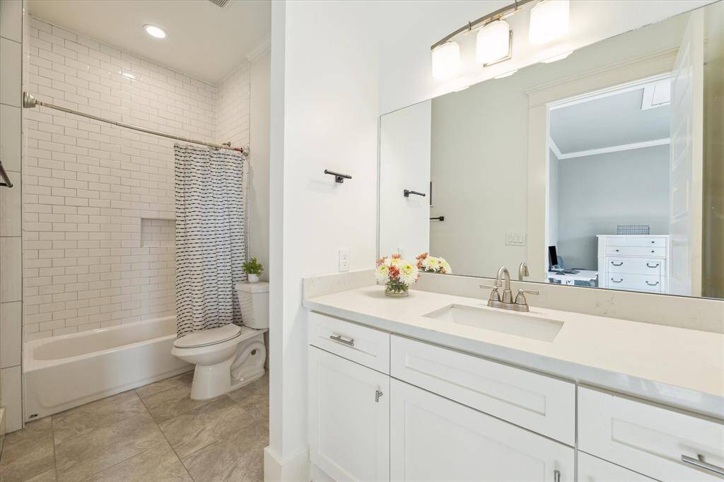 The en-suite bathroom features a modern design with a white vanity, a large mirror, and overhead lighting. It includes a bathtub with subway tile backsplash and a patterned shower curtain, complemented by light-color floor tiles for a fresh, clean look.