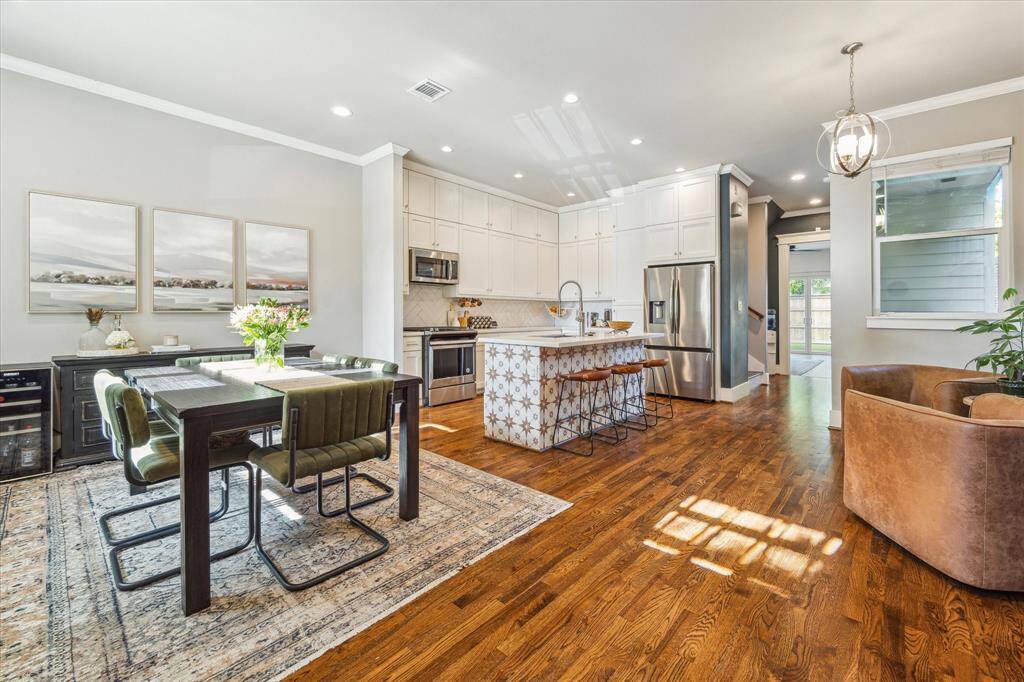 This photo showcases a bright, open-concept kitchen and dining area with hardwood floors. The kitchen features modern stainless steel appliances, white cabinetry, and an island with a patterned base and space for bar stools.