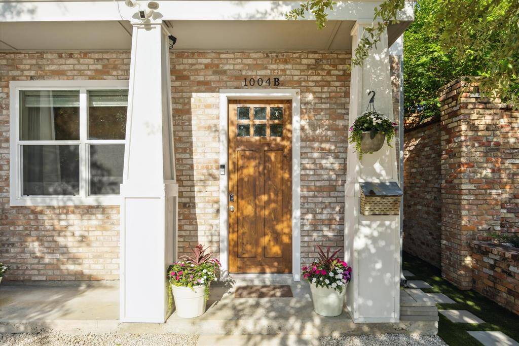 Charming brick exterior with a wooden front door, accented by a large window for natural light and potted plants. The inviting entryway is surrounded by greenery, enhancing the cozy appeal.