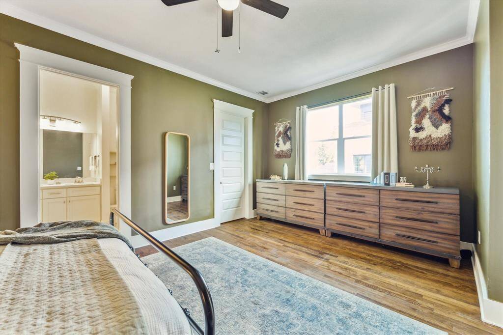 This spacious bedroom features warm green walls, hardwood flooring, and ample natural light. It includes a large window, space for sizable dresser, a stylish ceiling fan, and an ensuite bathroom with modern fixtures.