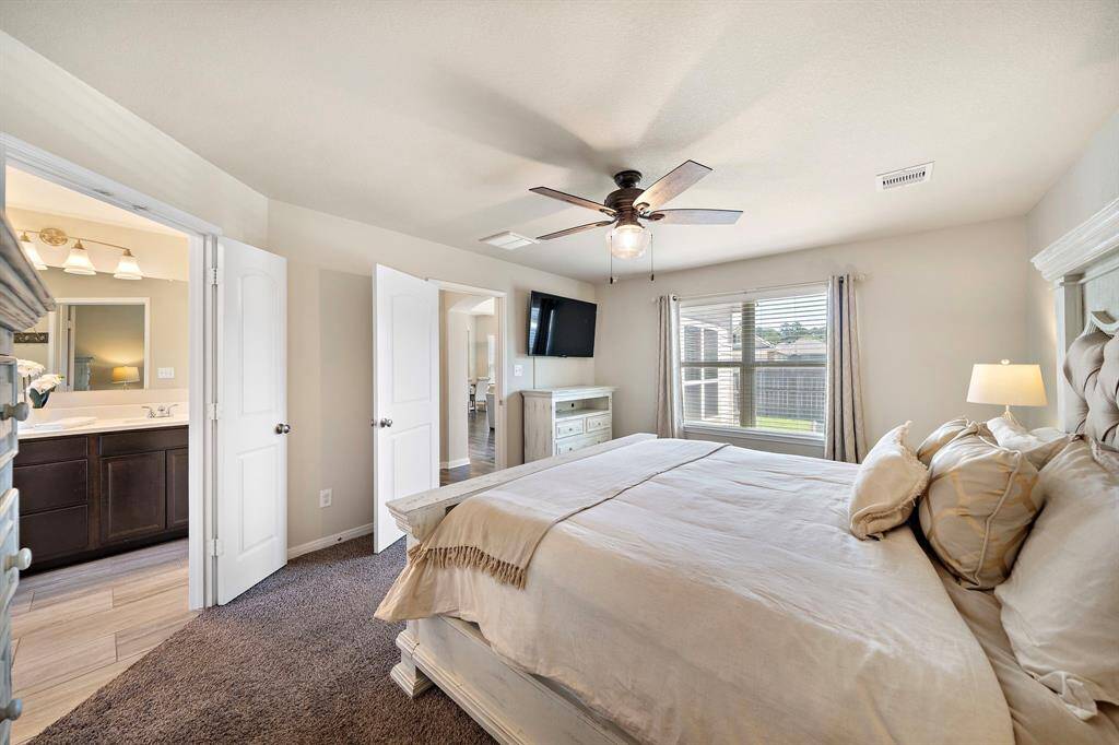 Bright primary bedroom suite features large windows with backyard views, plush carpeting, and a tray ceiling with an elegant ceiling fan. Direct ensuite bath access, neutral tones, and ample space for a king bed create a welcoming retreat designed for comfort and relaxation.