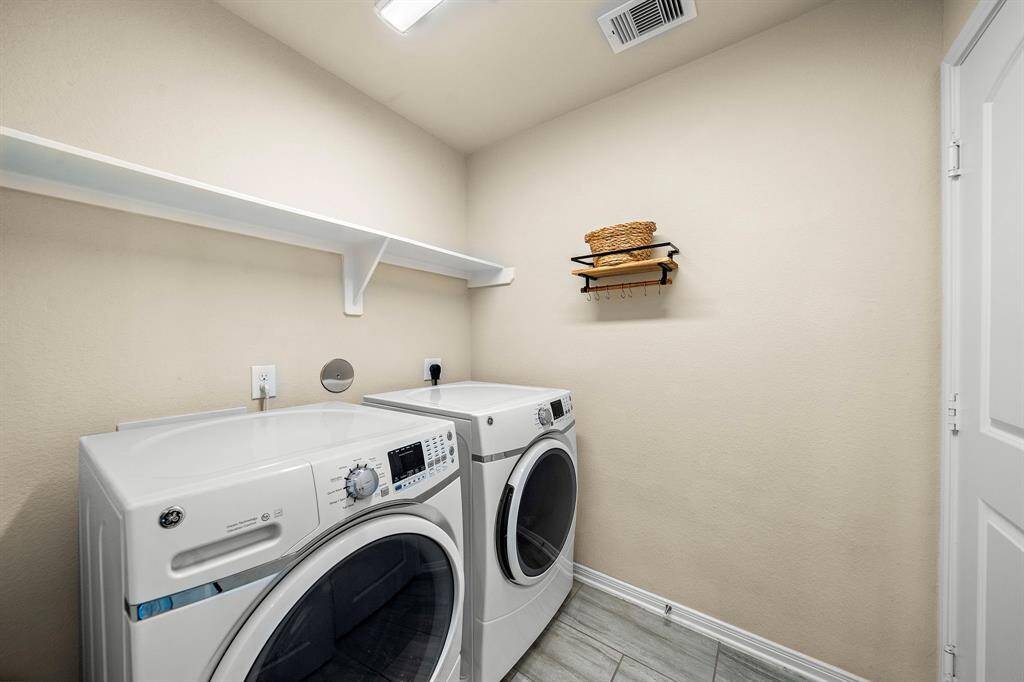 Dedicated laundry room includes a full-size washer and dryer, sleek tile flooring, and a built-in upper shelf for convenient organization. Wall hooks and shelving maximize storage, ensuring daily routines stay efficient and tidy.