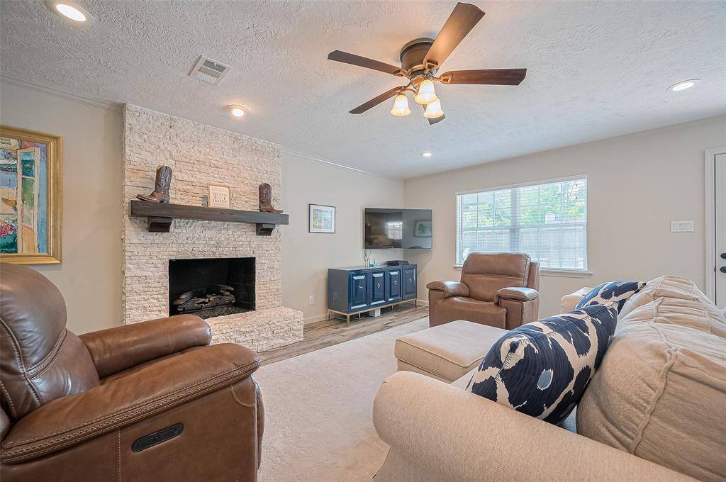 This spacious living room features a cozy stone fireplace, tile wood like flooring and ample natural light. It includes a ceiling fan with light fixture, crown molding, recessed lighting and large window, creating a welcoming atmosphere. A door leads outside, enhancing indoor-outdoor flow.