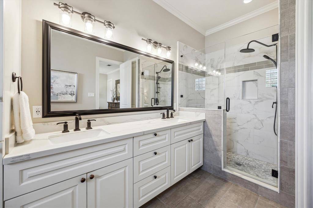 Enjoy the luxury of a frameless glass walk-in shower with rainfall showerhead and marble-look tile surround, paired with a spacious dual vanity and sleek black fixtures.