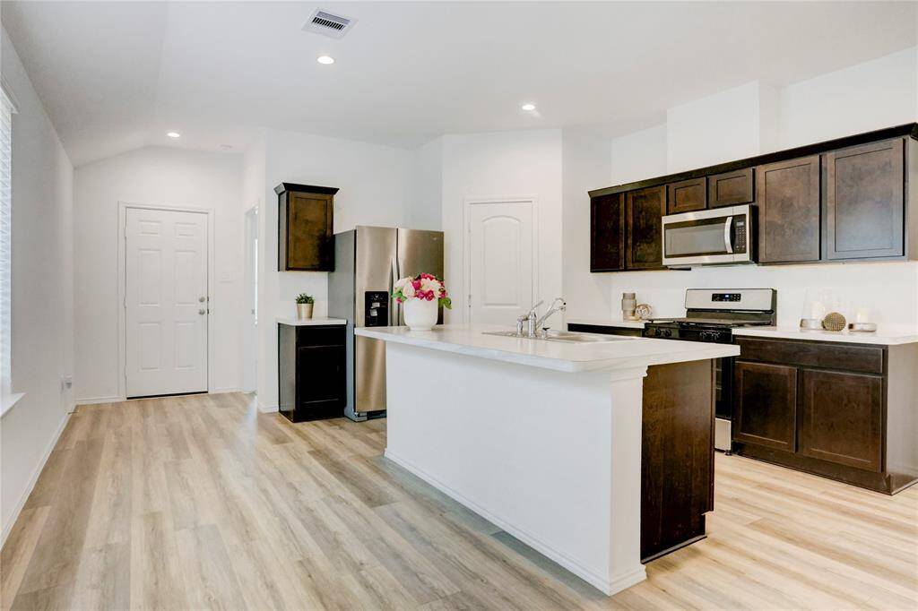 The kitchen offers abundant storage with upper and lower cabinetry. A central island with a built-in sink provides extra prep space and doubles as casual seating, all beneath recessed lighting and surrounded by large sunlit windows.