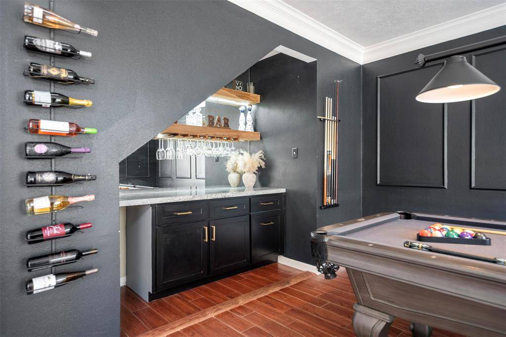 This inviting space features a stylish home bar with a sleek countertop, wine rack, and glass storage. Adjacent is a pool table, ideal for entertaining, set against rich wood flooring and elegant dark walls with modern paneling.