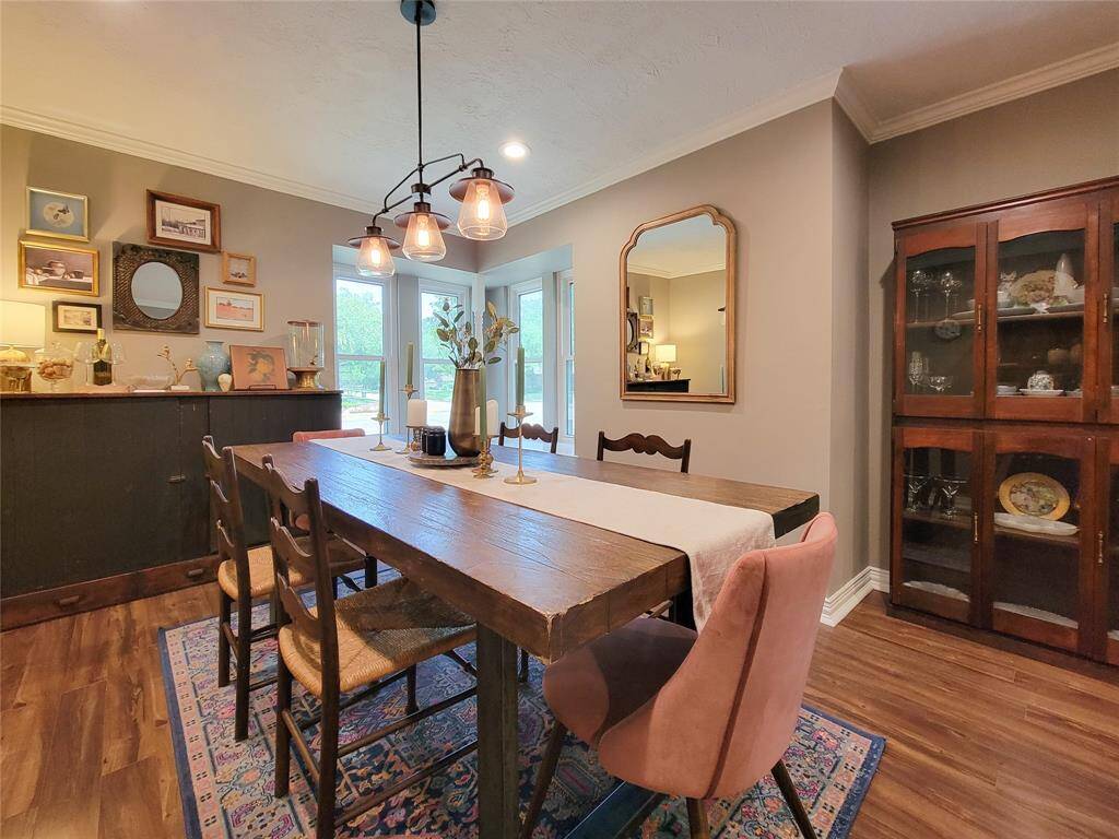 This dining room features a warm, inviting atmosphere with a rustic wooden table, eclectic chairs, and a stylish light fixture. There's a mix of modern and vintage decor with a gallery wall and a classic china cabinet. Large windows provide natural light, complementing the cozy, elegant vibe.