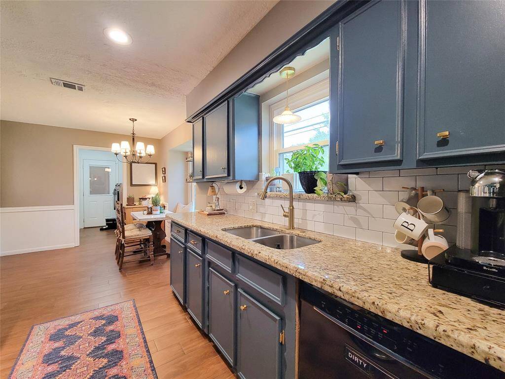 This kitchen features elegant blue cabinetry with brass hardware, granite countertops, and a double sink. It has a cozy dining area with a classic chandelier, wood-look tile floors, and a bright, welcoming atmosphere enhanced by a large window.