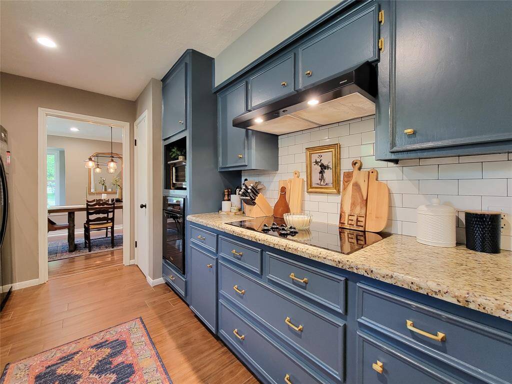 This kitchen features elegant blue cabinets with gold hardware, granite countertops, and a white subway tile backsplash. It opens to a dining area with stylish lighting, wood-look tile flooring, and ample natural light.
