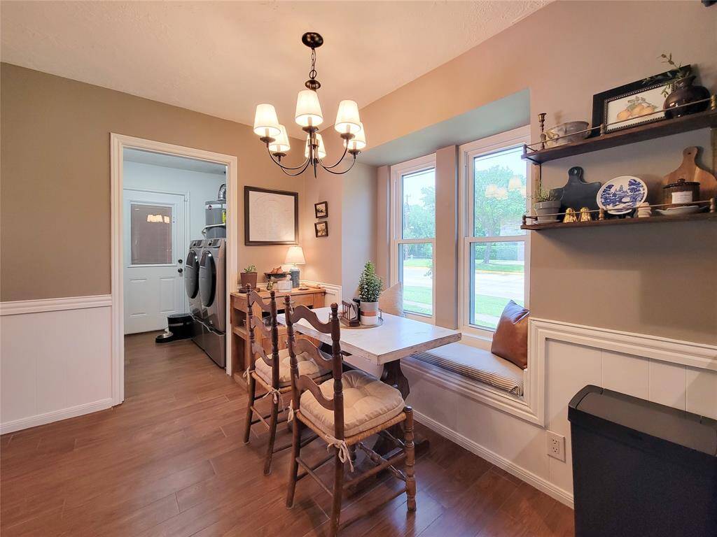 This cozy dining nook features a built-in window seat, wooden table, and chairs, illuminated by a charming chandelier. Adjacent is a laundry area with modern appliances, and shelves offer storage and display space. Large windows provide natural light and a view of the greenery outside.