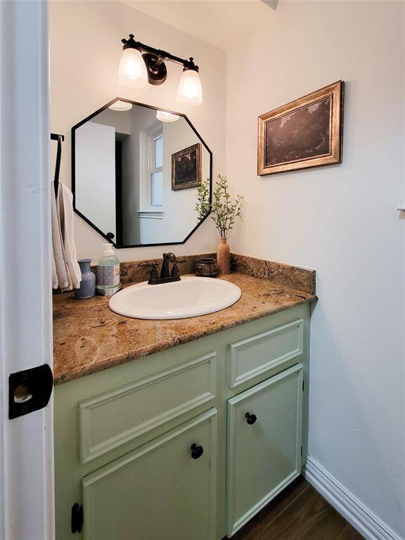 This photo showcases a powder bathroom with rustic charm. It features a light green vanity with a stone countertop, an oval sink, and black fixtures. The octagonal mirror and dual-light fixture add a modern touch, complemented by simple decor, including a small plant and framed art.