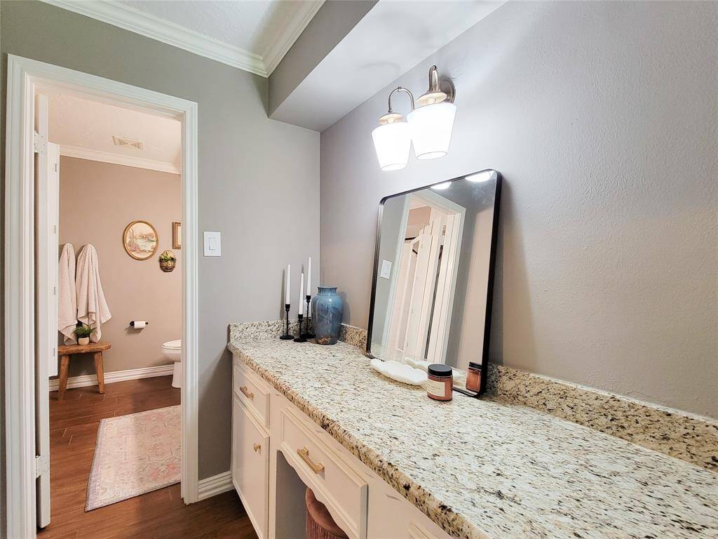 This photo showcases a stylish vanity area with granite countertops, modern light fixtures, and a large mirror. The space is complemented by wood-look tile floors and neutral wall colors, leading to a separate toilet area with decorative accents.