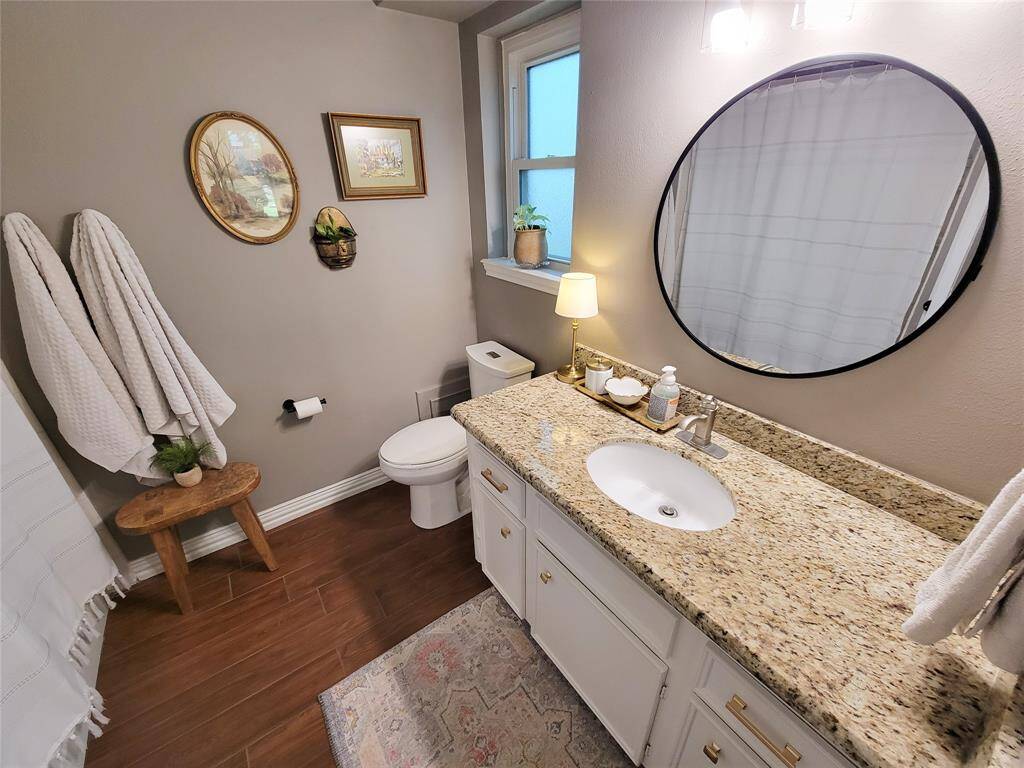 This bathroom features a granite countertop with a single sink and a round mirror above. It has a cozy, inviting feel with warm wood-look tile flooring, neutral walls, and tasteful decor. A window provides natural light, and there's a small plant adding a touch of greenery.