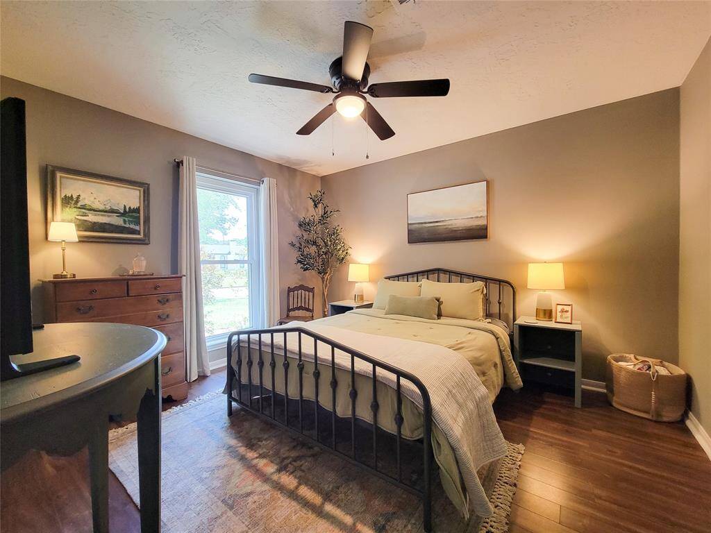 This cozy bedroom features warm, neutral tones with a modern metal bed frame. It has wood-like flooring, a ceiling fan, and large window for natural light. The room is accented with tasteful decor and functional furniture, creating a welcoming and serene atmosphere.