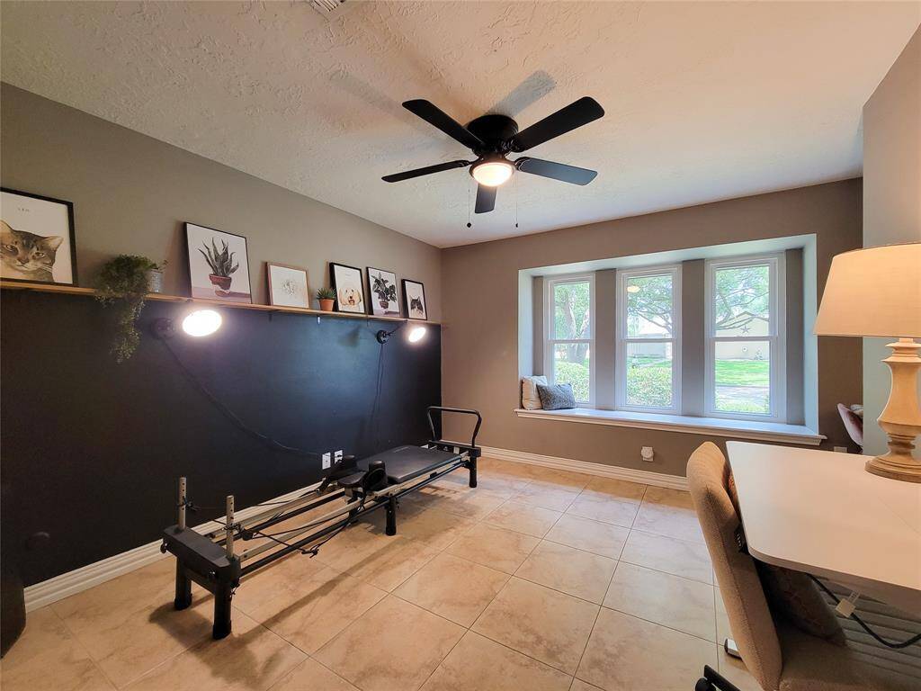 This room features a bright and airy space with a large window seat, perfect for relaxing. It includes a ceiling fan, tiled floors, and a versatile setup that can be used as a home office or exercise area. The decor is modern, with wall art and a shelf for additional storage or display.