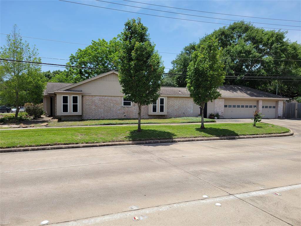 This is a single-story home with a light brick exterior and a three-car garage. It features a well-maintained lawn with a few trees and is situated on a corner lot. The house has large windows, adding to its curb appeal.