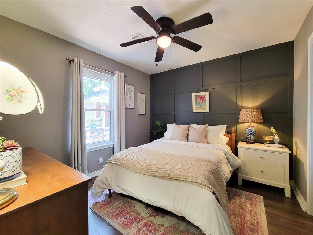 Cozy bedroom with a modern, dark accent wall and ceiling fan. Features a large window, natural light, and stylish decor. Includes a comfortable bed, nightstand, and dresser, creating a warm and inviting atmosphere.