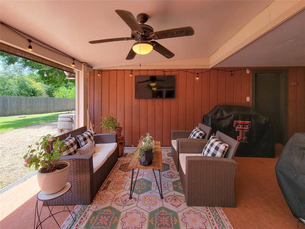 This outdoor patio features a ceiling fan, comfortable wicker seating with cushions, and a patterned rug. There's a mounted TV, string lights for ambiance, and a spacious view of the backyard, perfect for relaxing and entertaining.