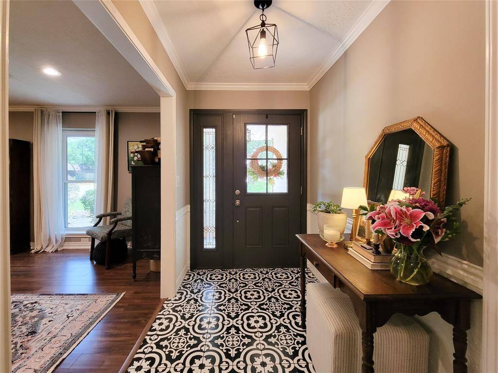This inviting entryway features a stylish black door with glass panels, patterned tile flooring, and warm lighting, leading into a cozy living space with wood-look tile floors and large windows.