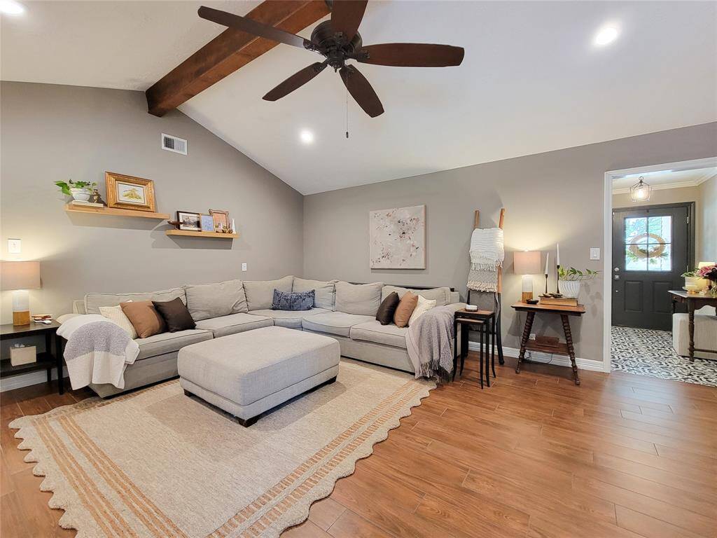 This inviting living room features a vaulted ceiling with a wooden beam and wood tile floors. It's beautifully lit with recessed lighting and natural light from a nearby entryway. The space is accented with cozy decor and a ceiling fan for comfort.