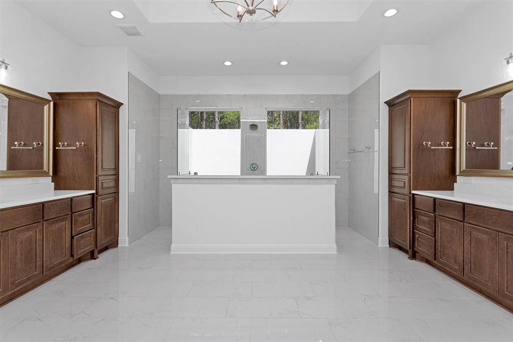 This luxurious spa-inspired bath features dual vanities, rich wood cabinetry, and an oversized walk-in shower framed by picture windows that bring the outdoors in with total privacy.