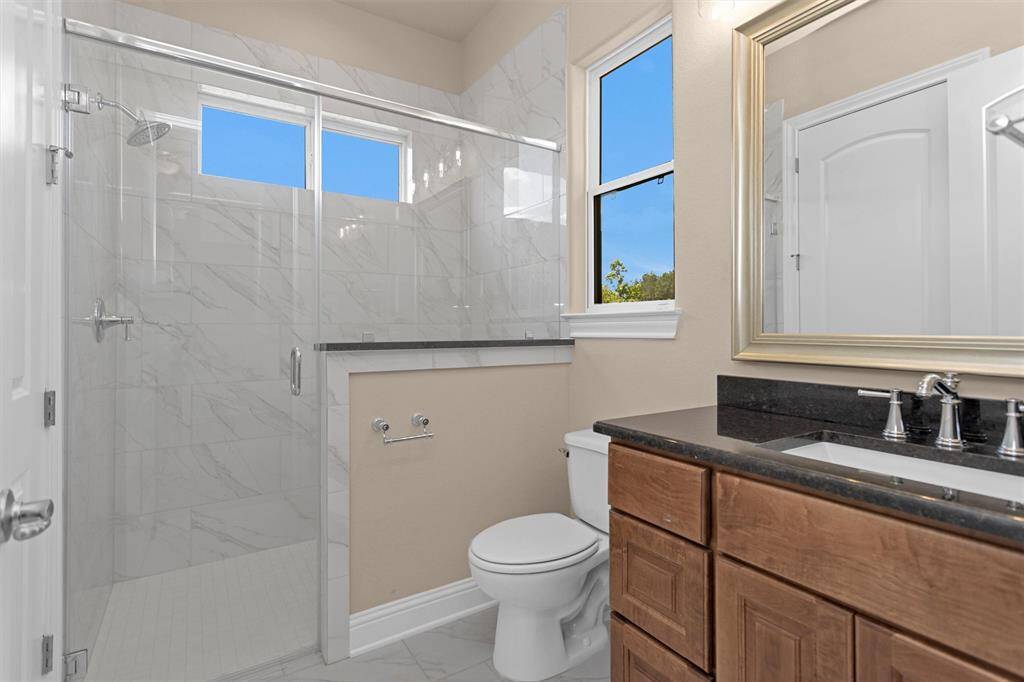 This en-suite bathroom features a spacious walk-in shower with elegant marble-style tile, natural light from dual windows, and warm wood cabinetry topped with sleek black granite counter.